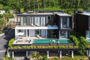 Modern two-story villa with floor-to-ceiling windows, a rooftop terrace, and a rectangular pool. Three people relax by the pool, surrounded by lush greenery and outdoor lounge areas.