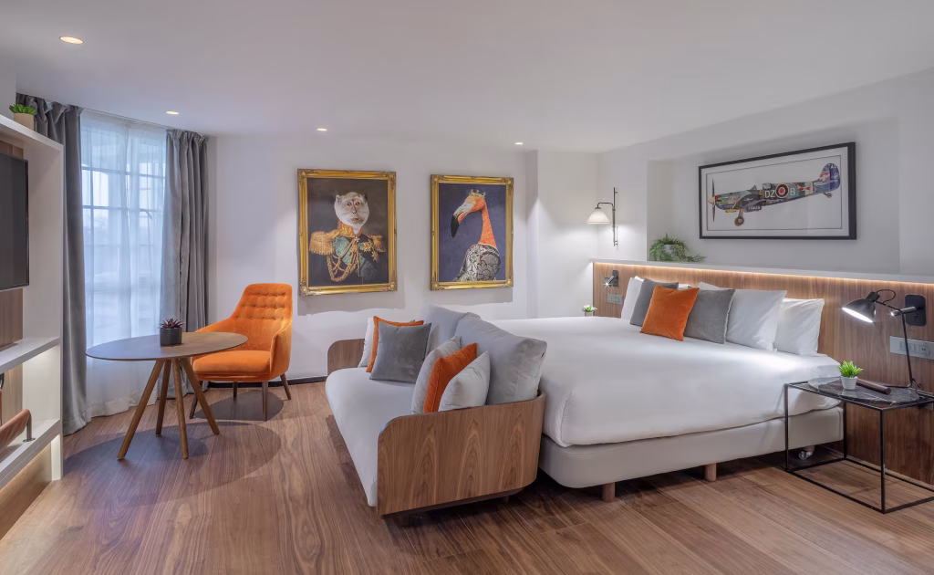 Modern hotel room with a large bed, orange and gray accents, a round table with an orange chair, wooden flooring, and whimsical animal portraits on the wall. Large window lets in natural light.