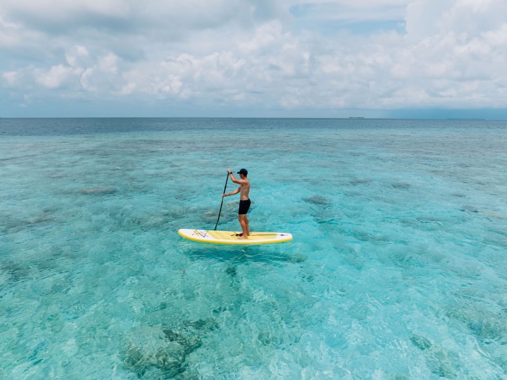 A person in swimwear paddleboards alone on clear, turquoise ocean water under a cloudy sky, surrounded by calm, shallow sea.