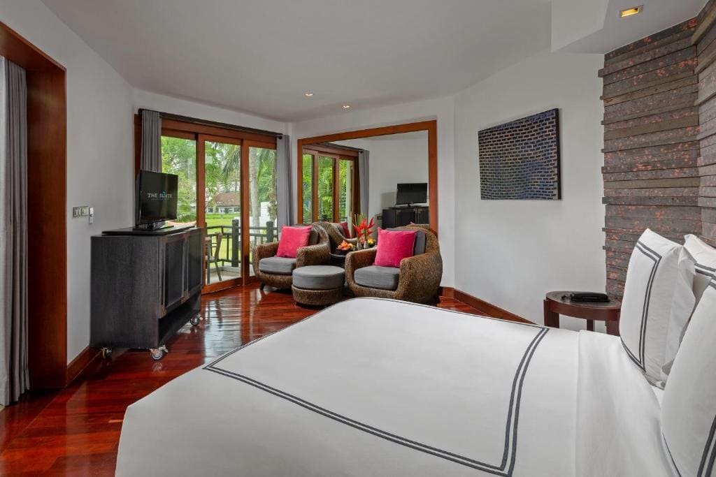 A bright hotel room with a large bed, two gray armchairs with pink cushions, a TV on a stand, and large windows opening to a view of greenery outside. The floor is polished wood.