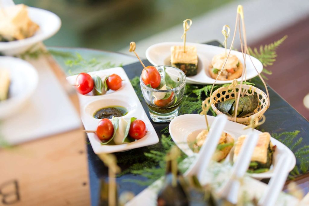 A close-up of an elegant appetizer spread featuring skewered cherry tomatoes with mozzarella, small bites wrapped in leaves, and various dipping sauces, all arranged on white plates with decorative greenery.