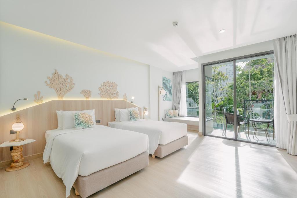 A bright, modern hotel room with two twin beds, light wood accents, ocean-themed wall decor, large windows, and a balcony with chairs overlooking greenery.