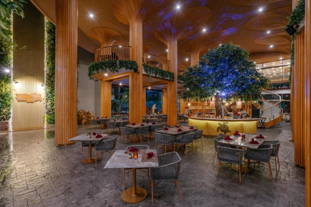 Elegant restaurant interior with tall wooden columns, tree-like ceiling structures, marble tables with woven chairs, a central tree, warm lighting, and greenery accents creating a cozy and upscale atmosphere.
