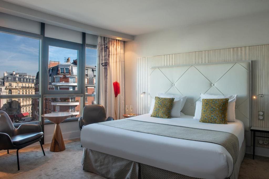 A modern hotel room with a large bed, two yellow-patterned pillows, a small table with two chairs by a window, and a view of city buildings outside under a blue sky.