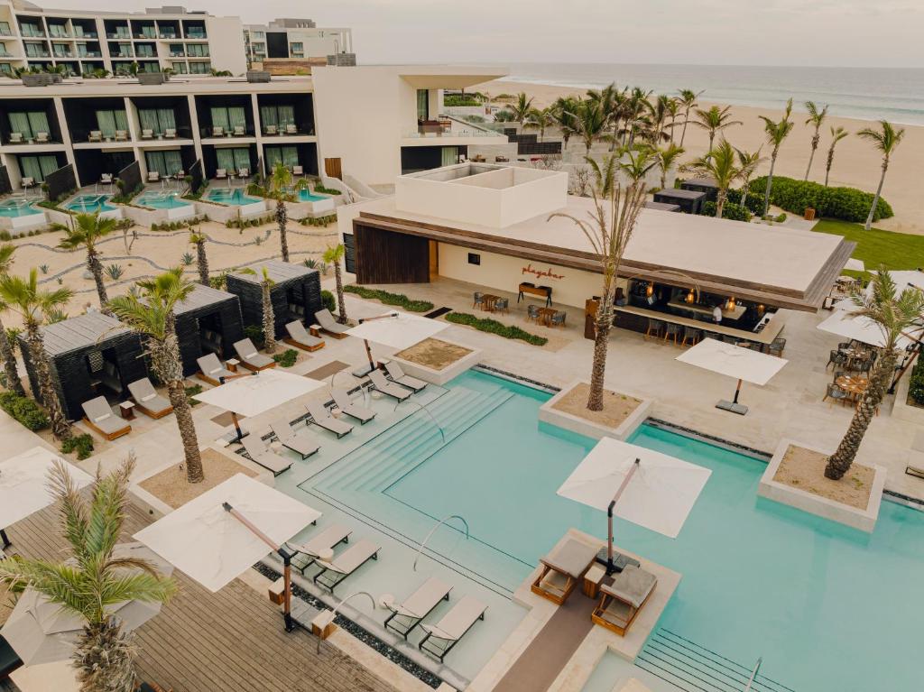 Aerial view of a modern resort with a large outdoor pool surrounded by lounge chairs, umbrellas, palm trees, cabanas, and a poolside bar. Multiple rooms with balconies and hot tubs face the pool area. The ocean is in the background.