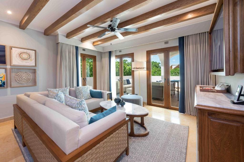 A modern living room with a beige sectional sofa, patterned pillows, two round coffee tables, large windows with curtains, wooden ceiling beams, and a ceiling fan. There is a balcony visible through glass doors.