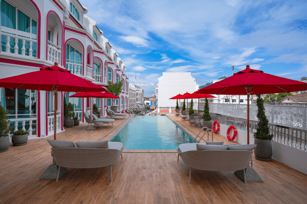 Outdoor hotel pool area with wooden deck, lounge chairs, and red umbrellas. Elegant building with white and pink accents lines the poolside under a partly cloudy blue sky.