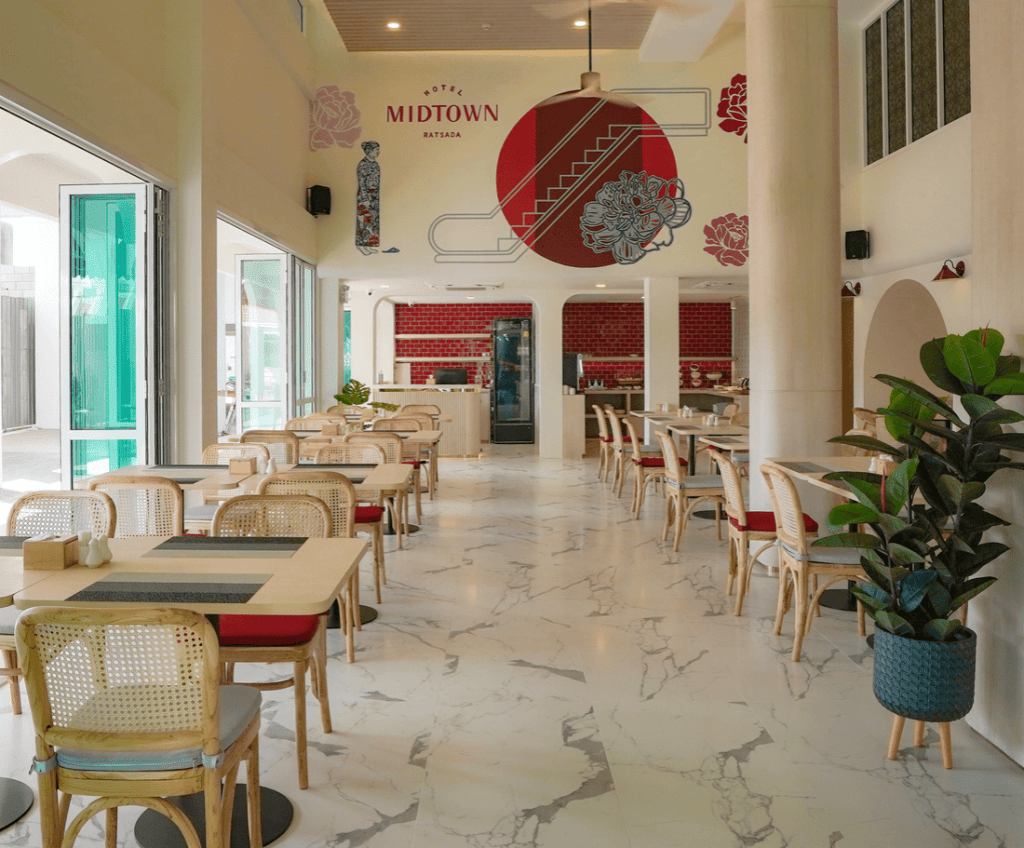 Bright, modern restaurant with marble floors, wooden tables and chairs, potted plants, and large windows. A mural with Hotel Midtown Batam and red accents decorates the back wall near the counter.