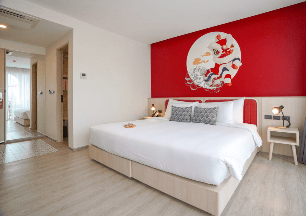 A modern hotel room with a large bed, white bedding, decorative pillows, nightstands with lamps, and a bold red accent wall featuring an artistic mural. There is natural light from a window and an open doorway to another room.
