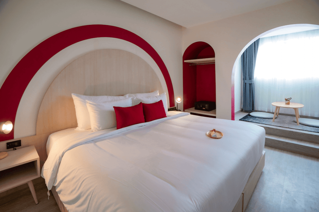 A modern hotel room with a large bed, white bedding, red accent pillows, and a red arch design on the wall. There are nightstands with lamps, an open wardrobe, and a sitting area near a window with sheer curtains.