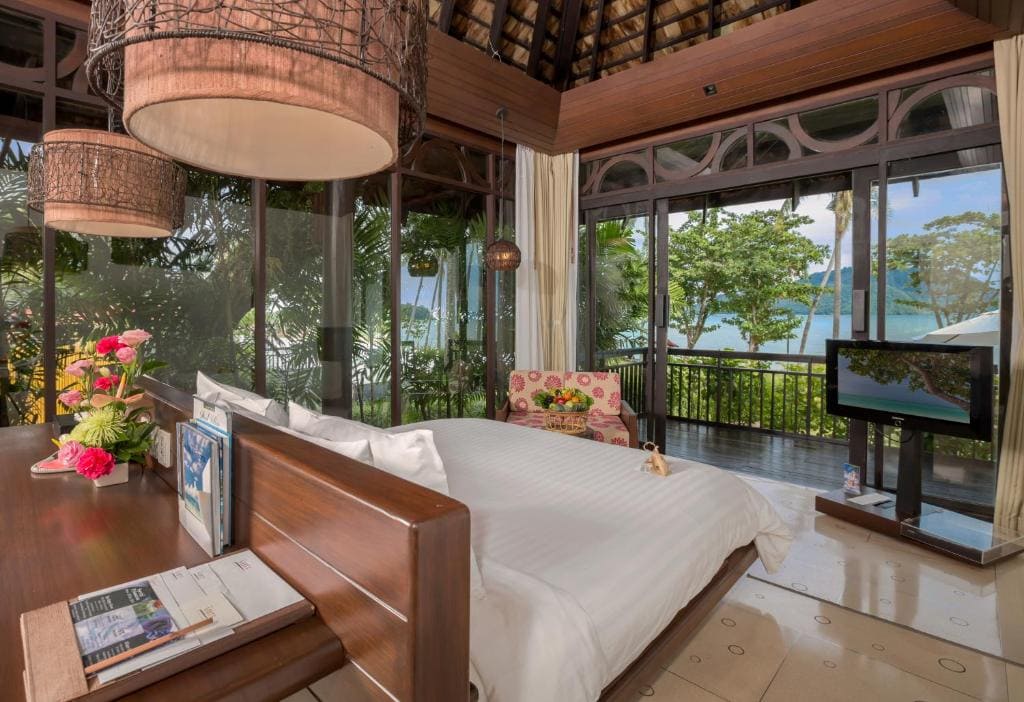 A cozy tropical hotel room with a large bed, fresh flowers, and a TV, featuring floor-to-ceiling windows offering views of lush greenery and a blue ocean beyond.