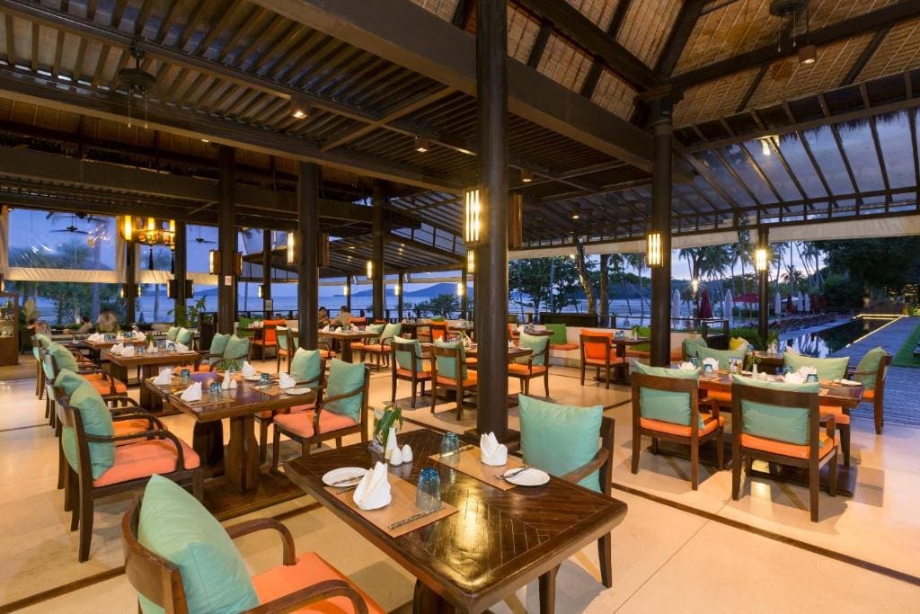 Open-air restaurant with wooden tables and cushioned chairs, set for dining. Warm lighting creates a cozy atmosphere. Large windows offer views of trees, outdoor pool, and evening sky in the background.