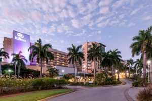 A large Hilton hotel with tall palm trees lining the curved driveway, set against a pastel sky with scattered clouds at sunset or sunrise. The building is brightly lit with a glowing Hilton sign.