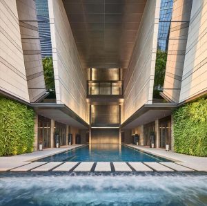 Modern building with tall, sleek walls and vertical gardens flanking a long, rectangular pool. Floating stone steps cross the pool, leading to a glass-walled structure at the end.