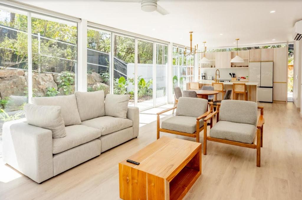 Bright, modern living room with light wood floors, a beige sofa, two matching armchairs, a wooden coffee table, and an open kitchen with dining area, large windows, and abundant natural light.