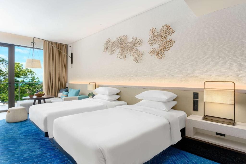 Modern hotel room with two neatly made twin beds, blue carpet, a beige accent wall with metal art, a side table with lamp, and a seating area by large windows with views of greenery outside.