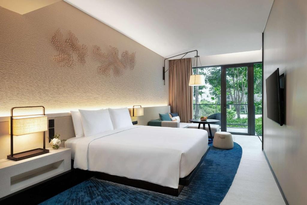 Modern hotel room with a neatly made bed, bedside lamps, a blue rug, a small seating area, a wall-mounted TV, large windows, and a view of greenery outside.
