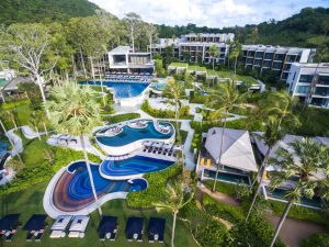 Aerial view of a modern resort featuring multiple pools with artistic patterns, lounge chairs, lush green landscaping, palm trees, and contemporary buildings surrounded by forested hills.