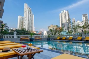 A rooftop swimming pool with yellow lounge chairs and side tables, two colorful drinks, and a hat. Tall modern buildings rise in the background under a clear blue sky.