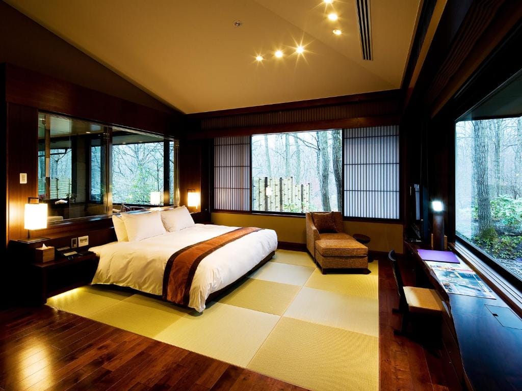A spacious, modern bedroom with a large bed, wood floors, tatami mats, a lounge chair, and floor-to-ceiling windows offering views of trees and natural light. Warm lighting creates a cozy atmosphere.