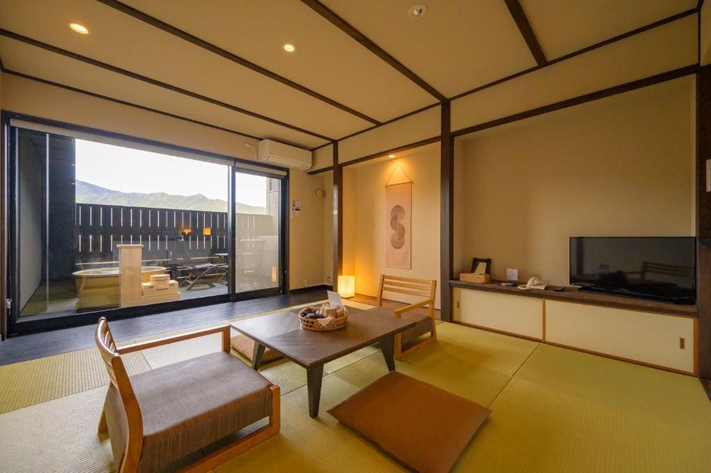 A traditional Japanese tatami room with low wooden table, floor cushions, and chairs. A TV sits on a cabinet, a scroll hangs on the wall, and large windows open to a balcony with mountain views.