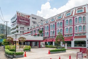 A mid-rise hotel named Midtown with white and maroon exterior, arched windows, and a red roof. The entrance has potted plants, a parking area with orange cones, and clear skies overhead.