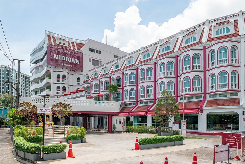 A mid-rise hotel named Midtown with white and maroon exterior, arched windows, and a red roof. The entrance has potted plants, a parking area with orange cones, and clear skies overhead.