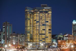 A tall, modern glass building labeled Park Hyatt is brightly lit at night, standing among other city buildings with busy traffic and streetlights visible in the foreground.