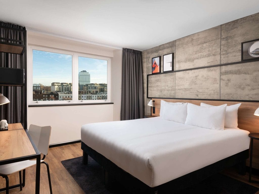 A modern hotel room with a large bed, white linens, bedside lamps, a wooden desk with a chair, wall art above the bed, and a window offering a city view with buildings under a blue sky.