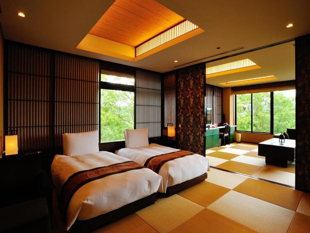 A spacious Japanese-style hotel room with twin beds, tatami mat flooring, wooden accents, soft lighting, large windows, and a scenic view of green trees outside. A low table and chairs are in the sitting area.