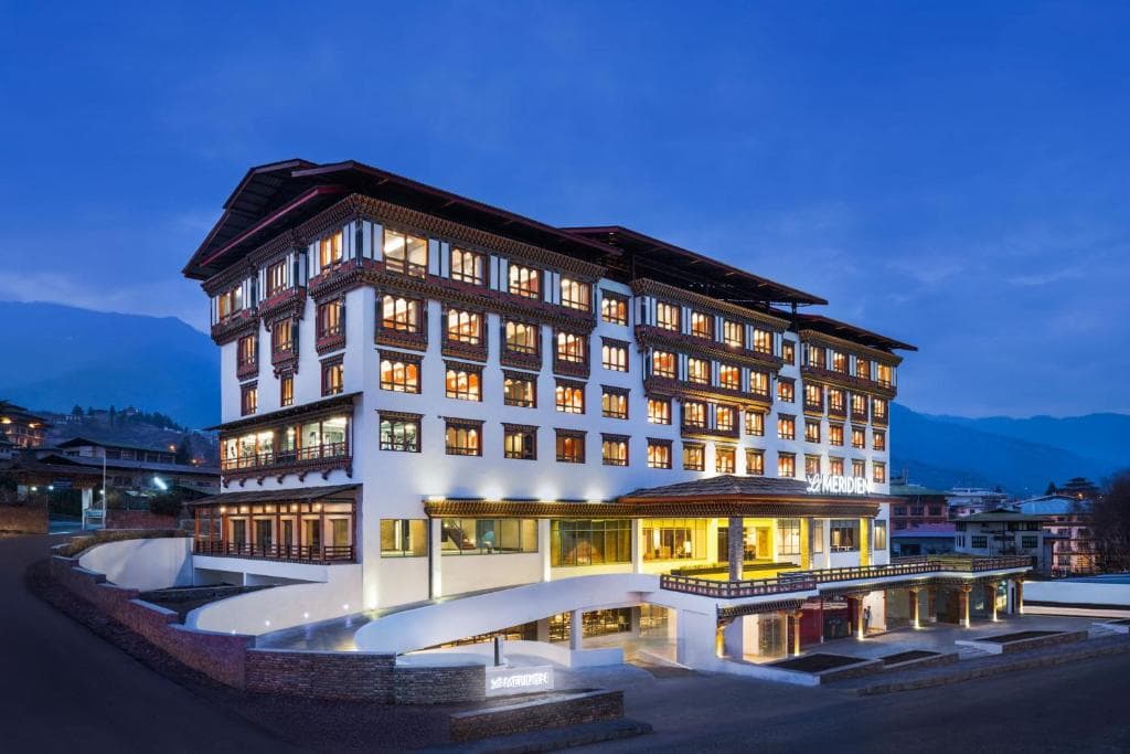 A large, multi-story hotel building with traditional Bhutanese architectural details is illuminated at dusk, with warm lights glowing from the windows and mountains visible in the background.