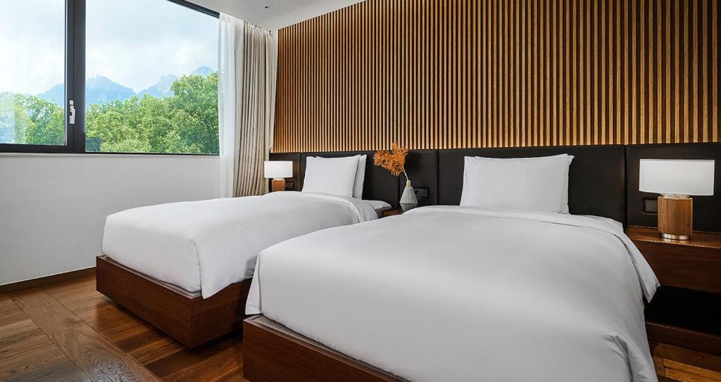A modern hotel room featuring two neatly made twin beds with white bedding, wooden floors, a large window with a view of trees and mountains, and a wall with vertical wooden slats behind the beds.
