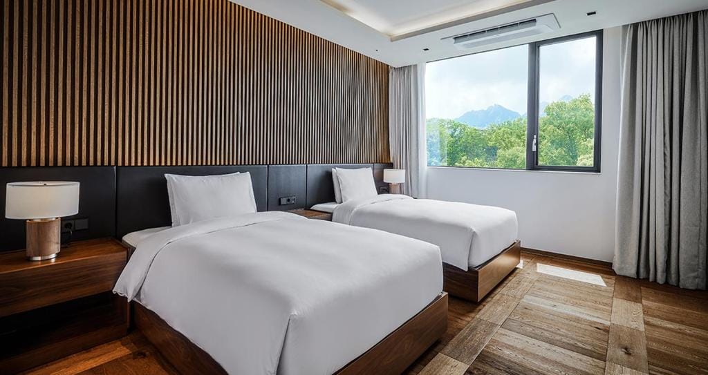 Modern hotel room with two single beds, wooden accents, a nightstand with a lamp, large window with a view of green trees and mountains, and light-colored curtains.