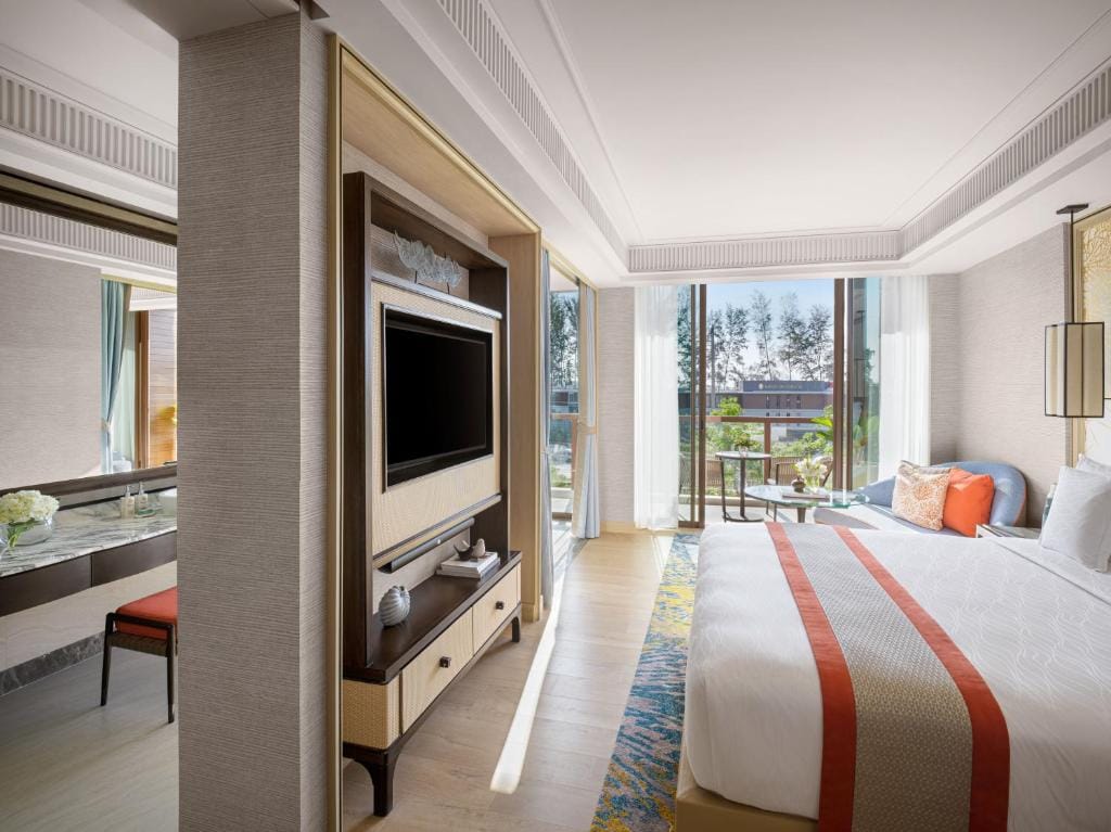 Modern hotel room with a large bed, flat-screen TV, decorative pillows, and floor-to-ceiling windows overlooking trees and a balcony. Light décor with wood and soft colors creates a bright, inviting atmosphere.