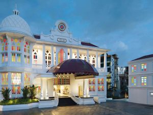 A grand white building with colorful stained-glass windows, pillars, and a domed tower, illuminated at dusk with a maroon entrance canopy and ornate architectural details.