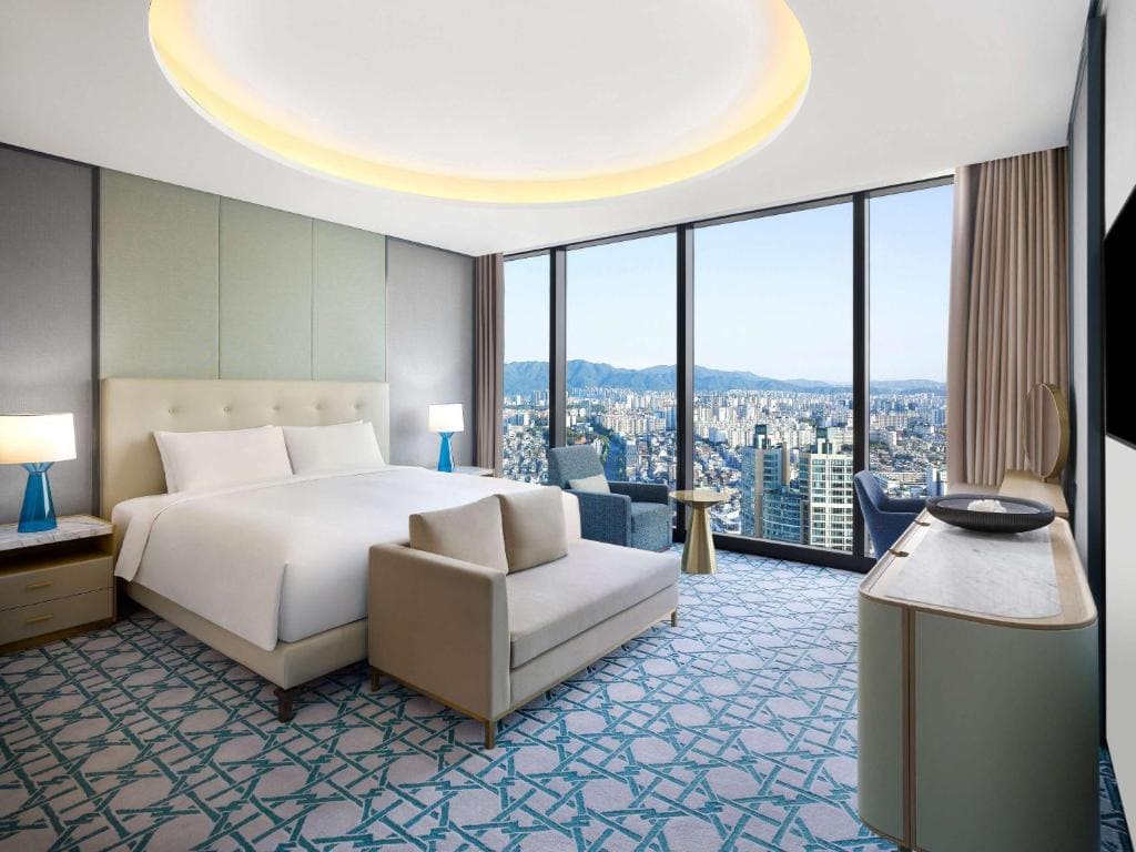 A modern hotel room with a large bed, stylish furniture, light blue patterned carpet, and floor-to-ceiling windows offering a panoramic city view under a bright, circular ceiling light.