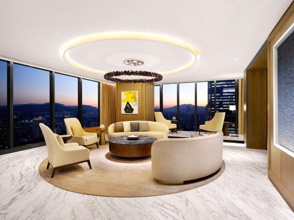 Modern, luxurious lounge with cream-colored sofas and chairs arranged around a circular table, large floor-to-ceiling windows showcasing a cityscape at sunset, and a statement ceiling light above.