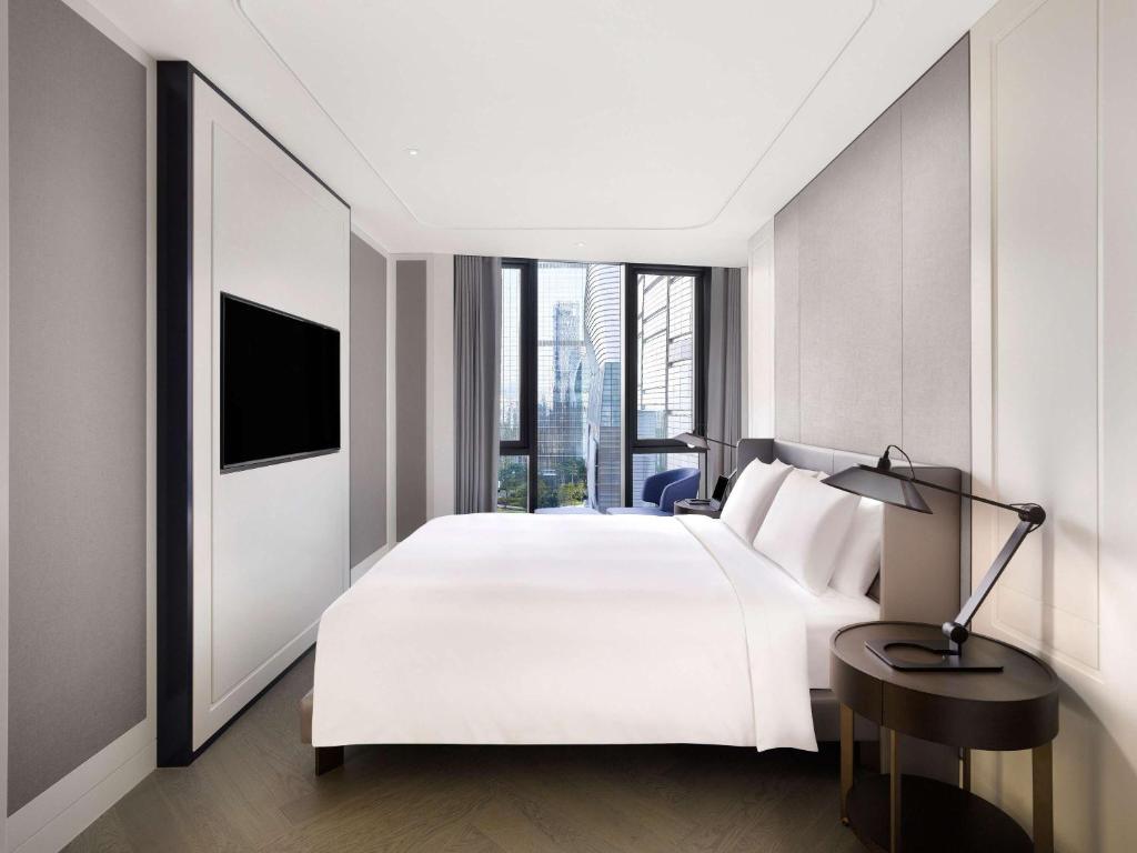A modern hotel room with a large bed, white linens, round bedside table with a lamp, wall-mounted TV, and floor-to-ceiling windows offering a city view.