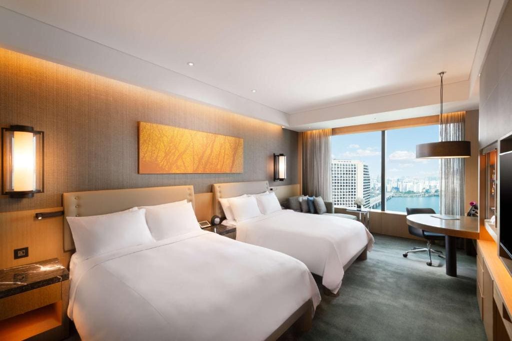 Modern hotel room with two large beds, a wall-mounted painting, bedside lamps, a desk and chair by a window with city skyline views, and soft, neutral décor.