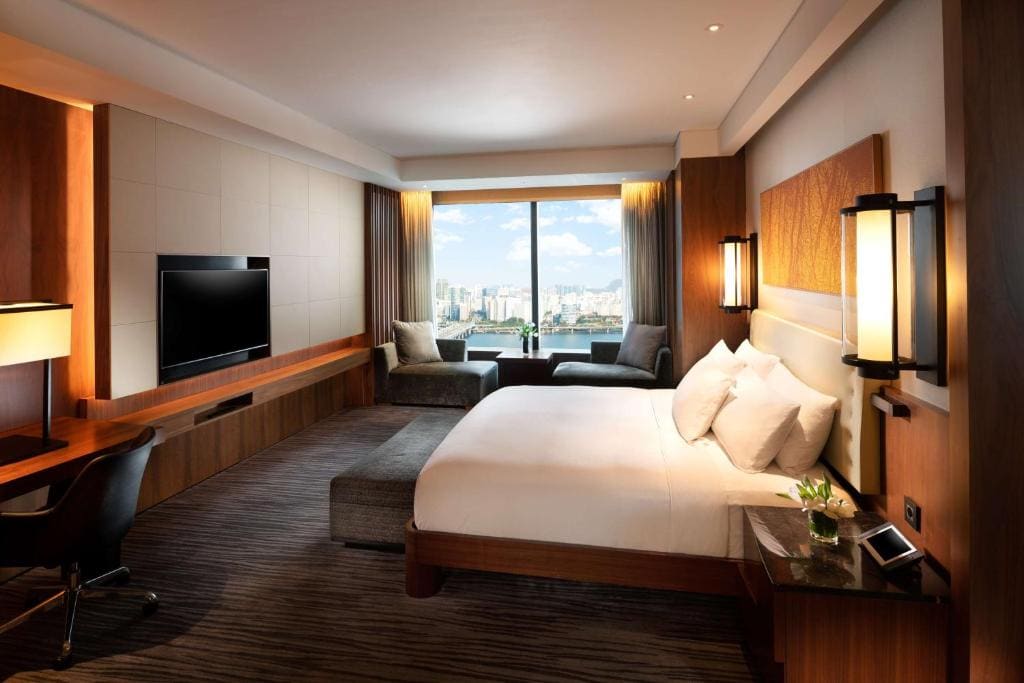 Modern hotel room with a large bed, desk, flat-screen TV, and a seating area by a window offering a city view. Warm lighting and neutral tones create a cozy, inviting atmosphere.