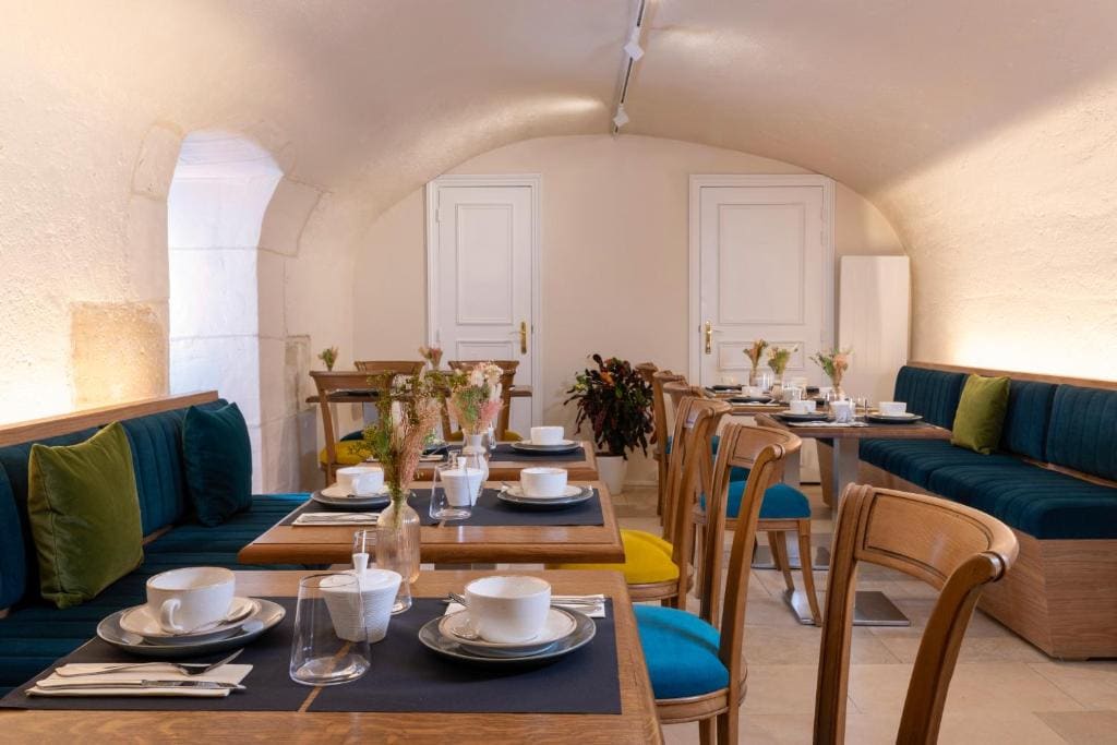 A cozy, modern café with wooden tables and chairs, blue cushioned benches, green and yellow pillows, neatly set with white dishes and glassware, and soft lighting under a vaulted ceiling.