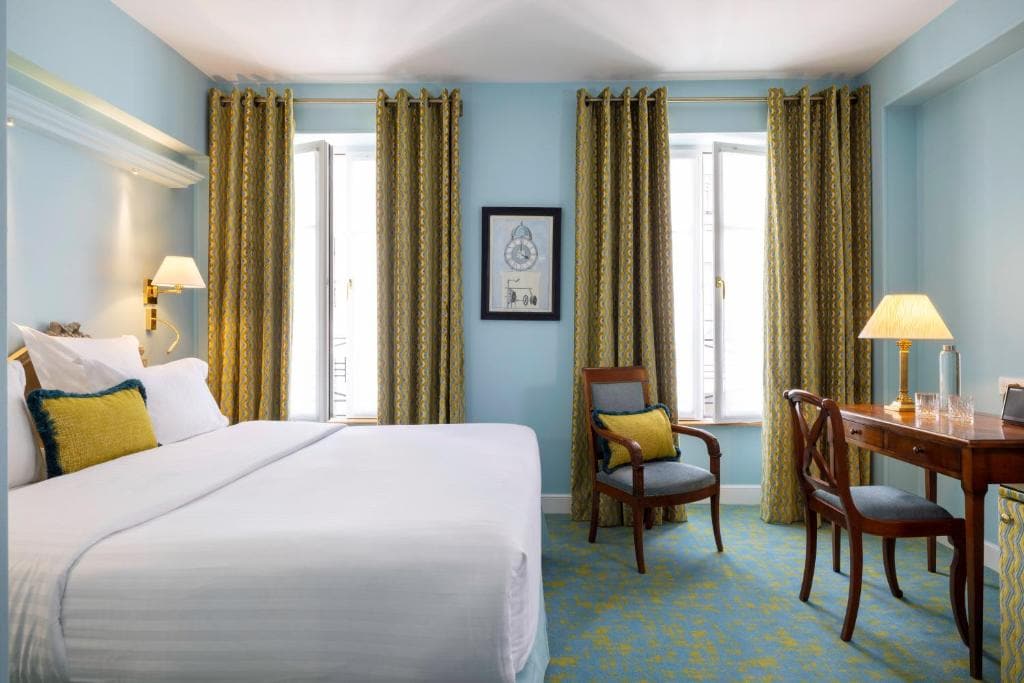 A bright hotel room with a large bed, yellow and blue cushions, two windows with gold curtains, a framed picture on a blue wall, a wooden desk with a lamp, and two wooden chairs on a patterned carpet.