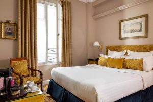 A cozy hotel room with a neatly made double bed, gold and white pillows, elegant curtains, a window, framed art on beige walls, a wooden chair, and a small table set with a cup, saucer, and coffee machine.
