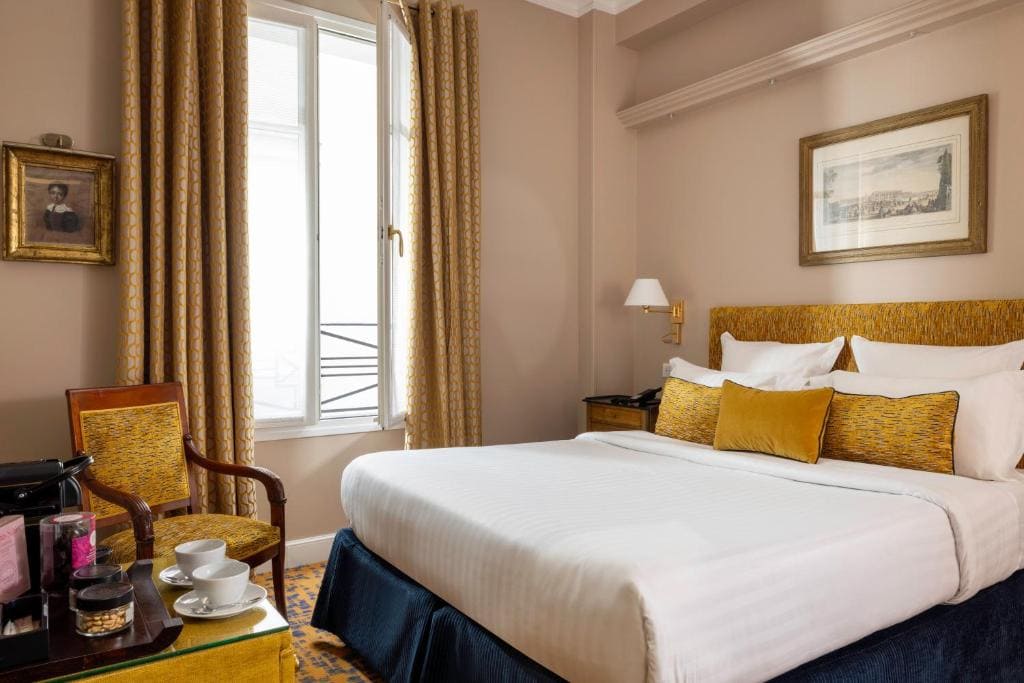A cozy hotel room with a neatly made double bed, gold and white pillows, elegant curtains, a window, framed art on beige walls, a wooden chair, and a small table set with a cup, saucer, and coffee machine.