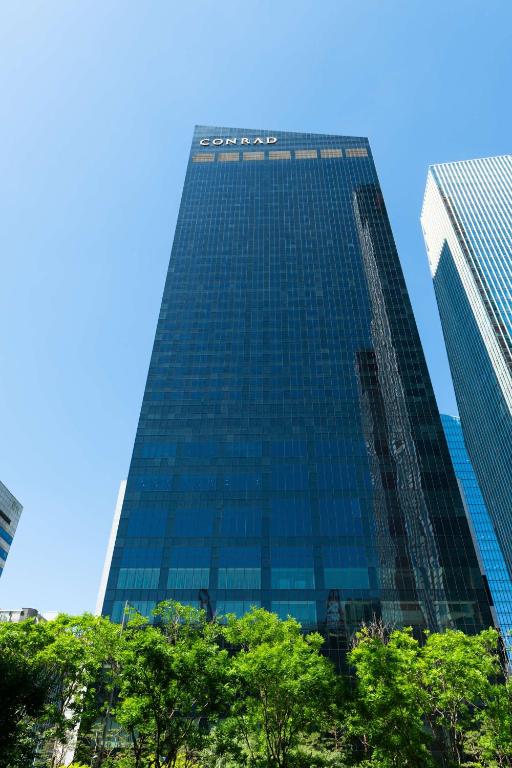 A tall, modern glass skyscraper with the word CONRAD at the top, set against a clear blue sky, with green trees in the foreground.