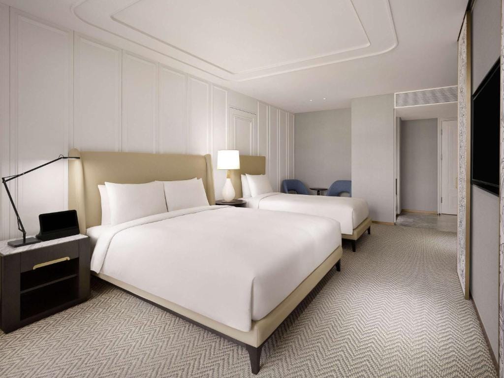 A modern hotel room with two large beds, white bedding, a nightstand with a lamp and desk, light-colored walls, carpeted floor, and a small seating area with two blue chairs in the background.