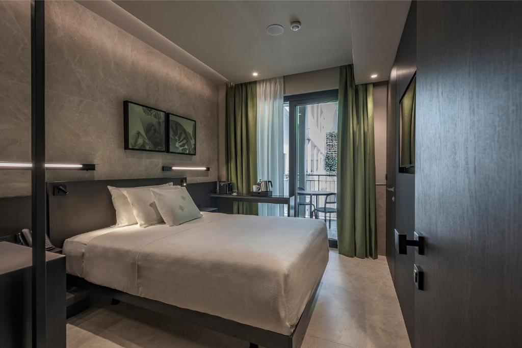 A modern hotel room with a double bed, neutral decor, two framed pictures above the bed, bedside tables, green and white curtains, and a glass door leading to a small balcony with chairs.