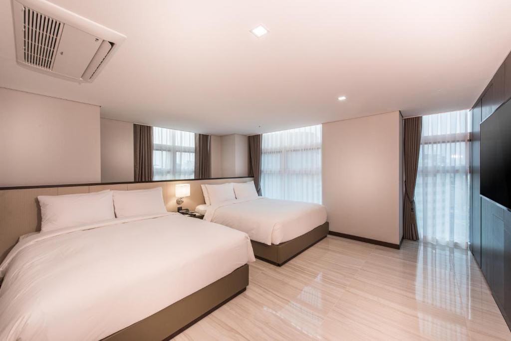 Minimalist hotel room with two large beds, white bedding, bedside table with lamp, light-colored tile floor, large windows with sheer curtains, and a wall-mounted TV. Room is well-lit and spacious.