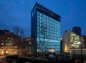 A modern office building with a grid of bright blue illuminated windows stands tall at dusk, surrounded by other buildings and leafless trees under a darkening sky.