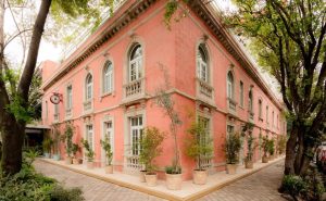 A large, elegant pink building with arched windows, white trim, and potted plants lining the sidewalk, shaded by tall trees, on a sunny day.